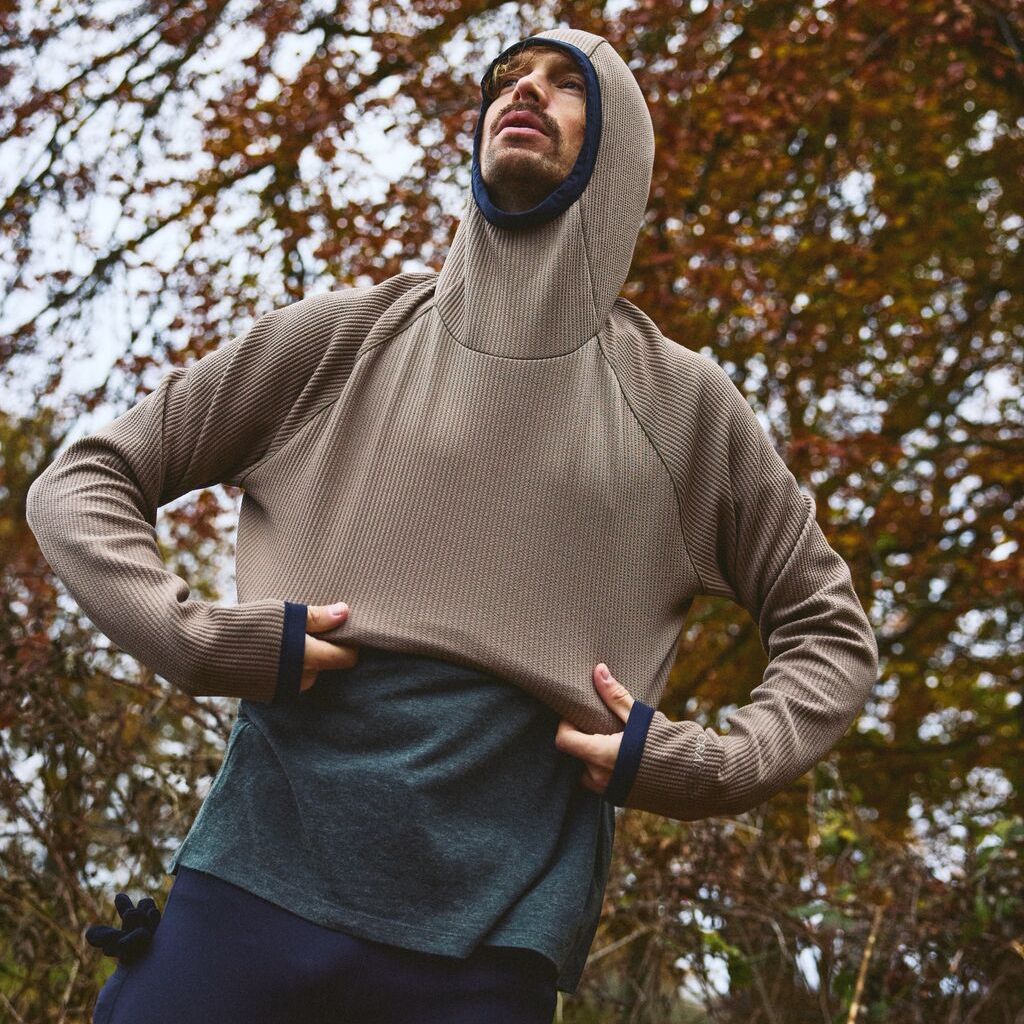 Person utendørs som justerer en brun hettegenser over en grønn skjorte, med høsttrær i bakgrunnen.