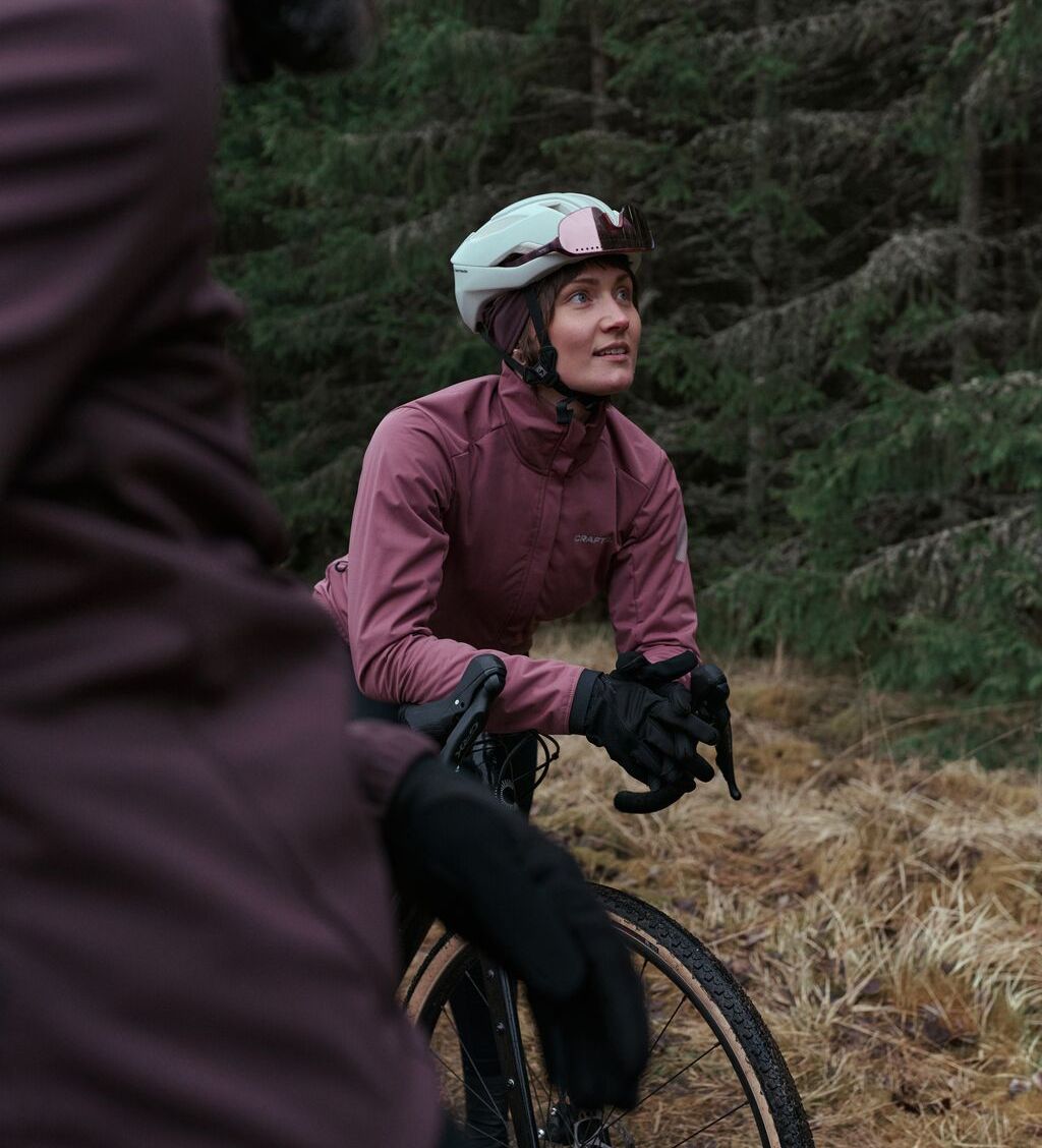 En syklist i lilla jakke og blå hjelm hviler på en sykkel i en skog og ser oppover. En annen syklist er delvis synlig i forgrunnen.