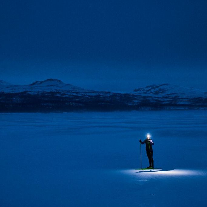 Skiløper med hodelykt på en snødekt slette om natten, omgitt av mørkeblå farger og fjerne snødekte fjell.