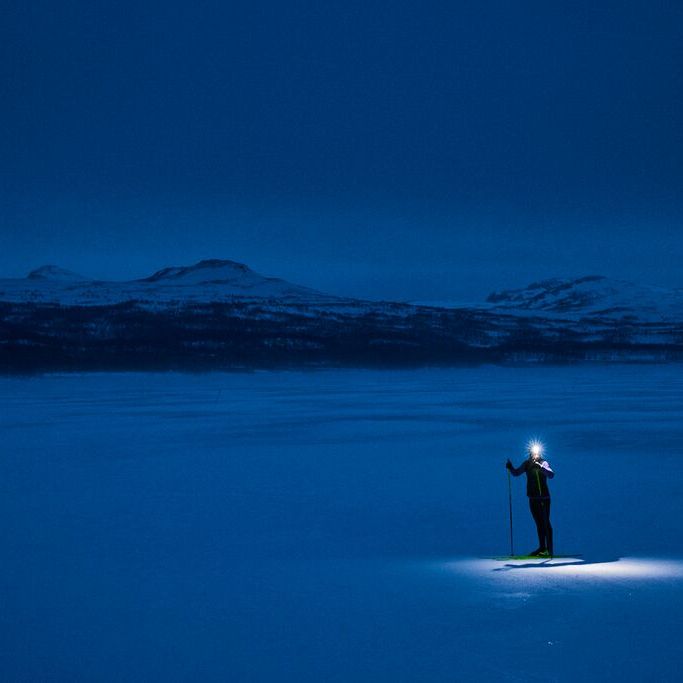 Skiløper med hodelykt på en snødekt slette om natten, omgitt av mørkeblå farger og fjerne snødekte fjell.