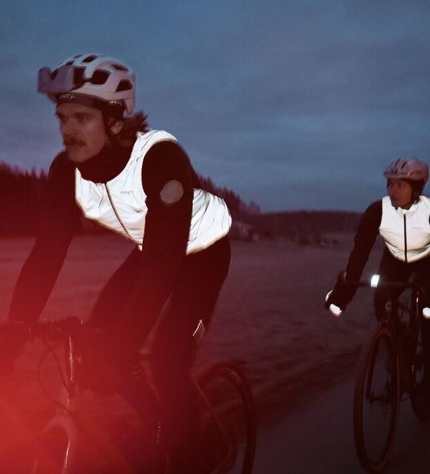 To syklister med hjelm og refleksvester sykler på en vei i skumringen, med trær i bakgrunnen og et rødt lysbluss i forgrunnen.