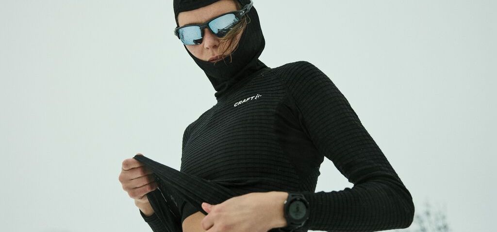 Person i svart vintersportsklær og reflekterende solbriller, som justerer toppen sin i et snødekt landskap.