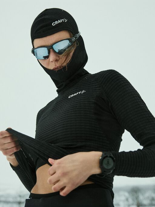 Person i svart vintersportsklær og reflekterende solbriller, som justerer toppen sin i et snødekt landskap.
