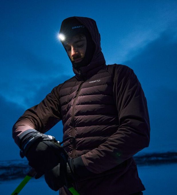 Person iført vinterutstyr og hodelykt, som justerer skistaver i et snødekt landskap i skumringen.