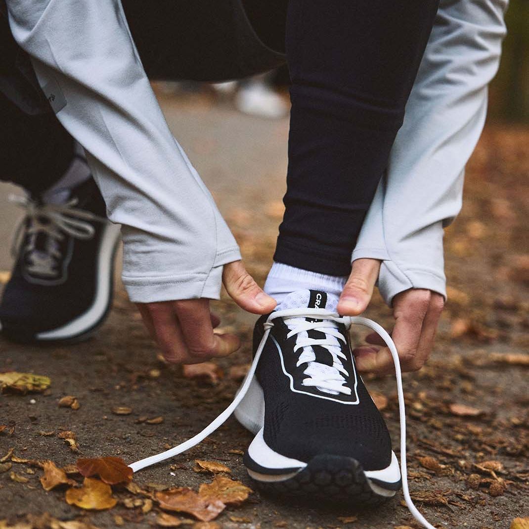 En person som knytter skolissene på et par svarte joggesko på en sti dekket av løv, kledd i svarte leggings og en grå jakke.