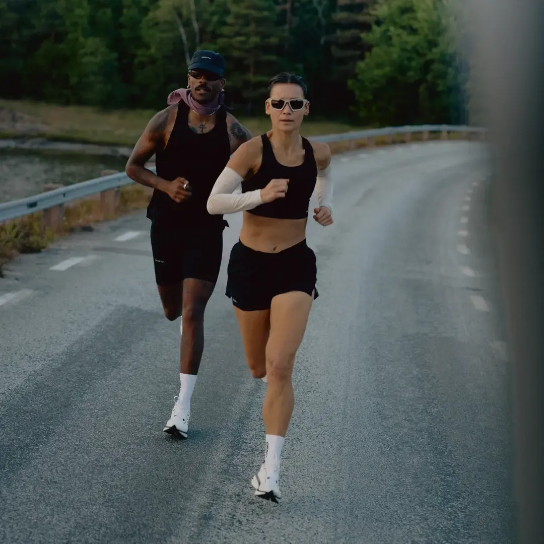 To løpere i treningsklær som jogger på en landevei, omgitt av grøntområder, med fokus på den forreste løperen som har på seg solbriller.