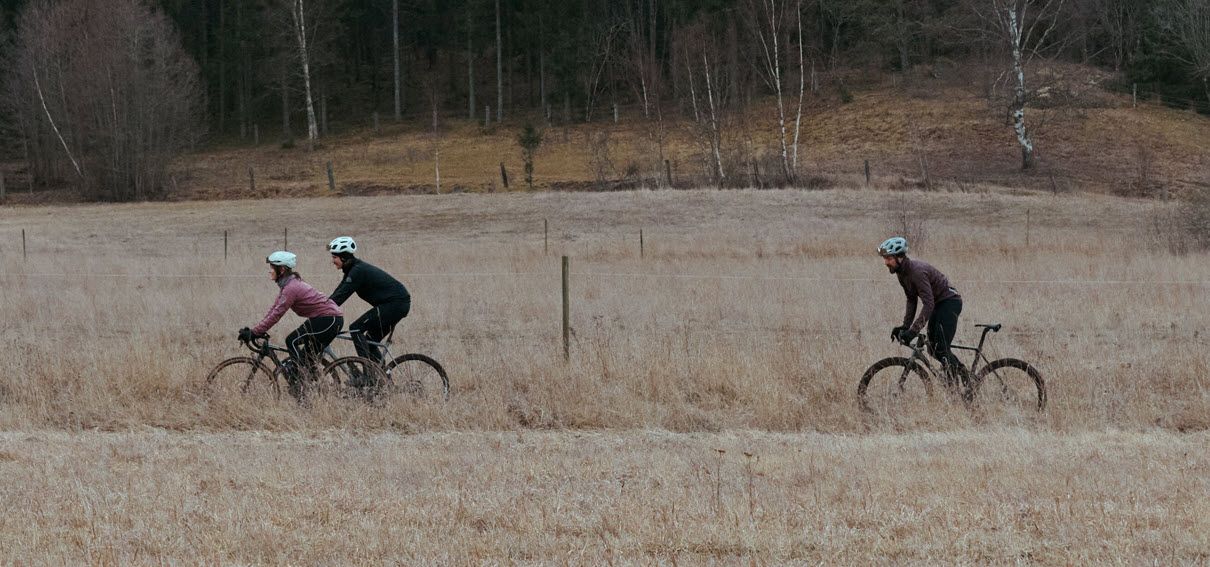 Tre syklister sykler på en åpen eng med skog i bakgrunnen på en overskyet dag.