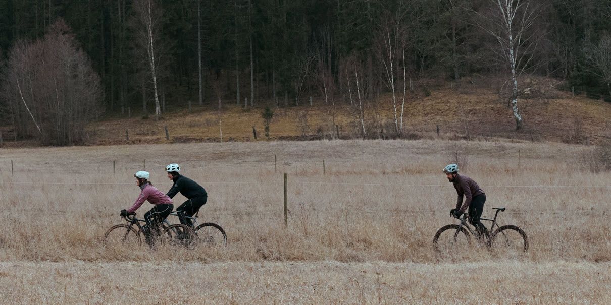 Tre syklister sykler på en åpen eng med skog i bakgrunnen på en overskyet dag.