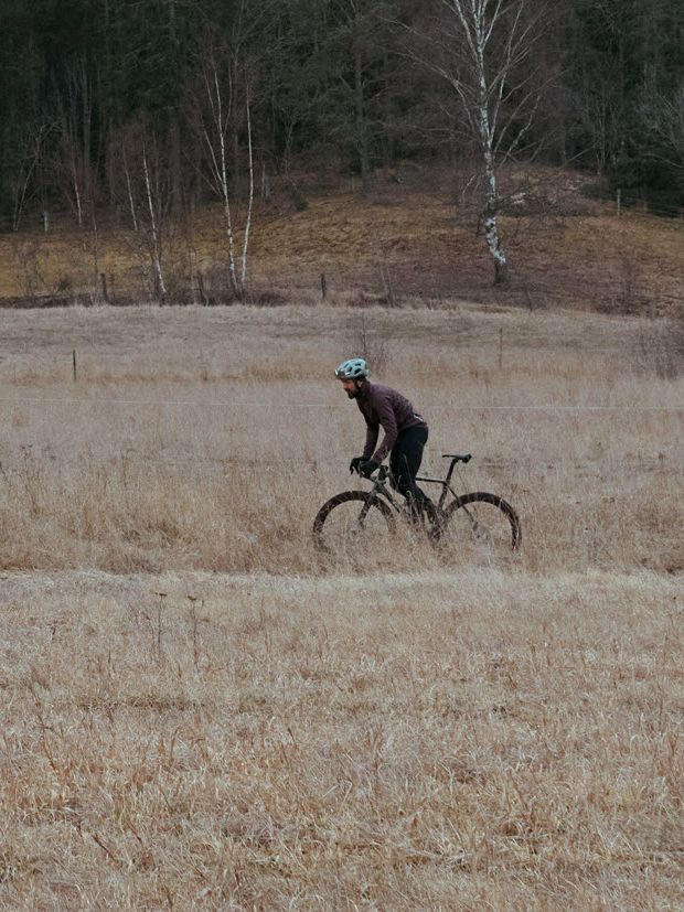 Tre syklister sykler på en åpen eng med skog i bakgrunnen på en overskyet dag.