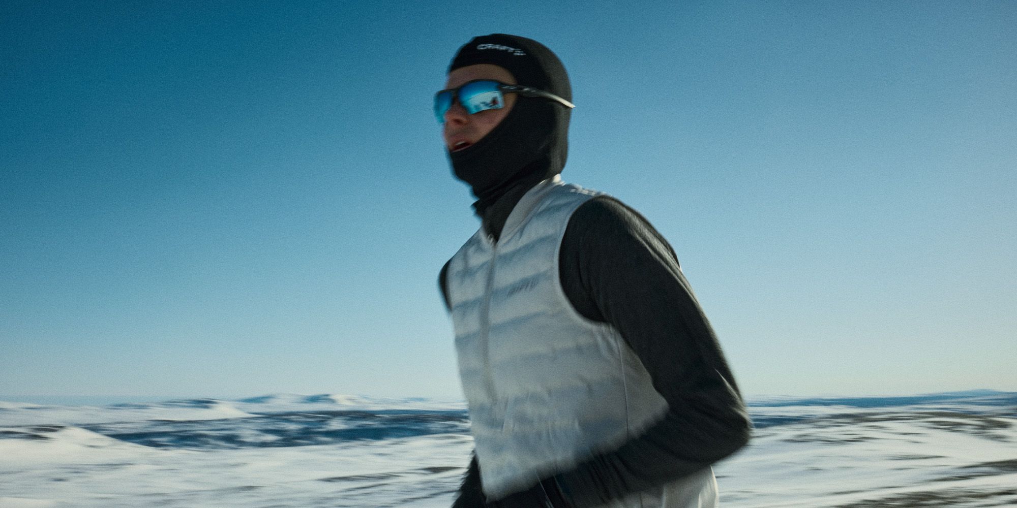Person som jogger i snødekt landskap iført vinterklær, inkludert en hvit vest, svart balaklava og reflekterende solbriller under en klar blå himmel.