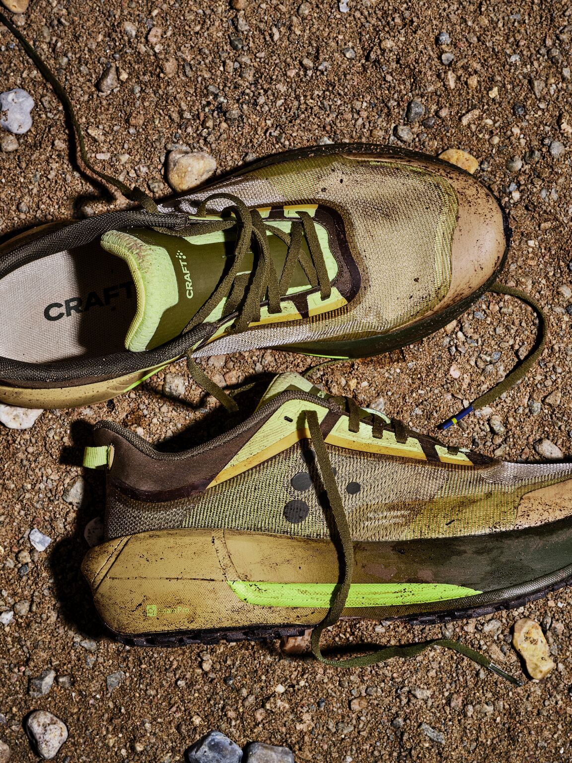 Slitte joggesko med grønne detaljer på en grusete jordoverflate, omgitt av små steiner.