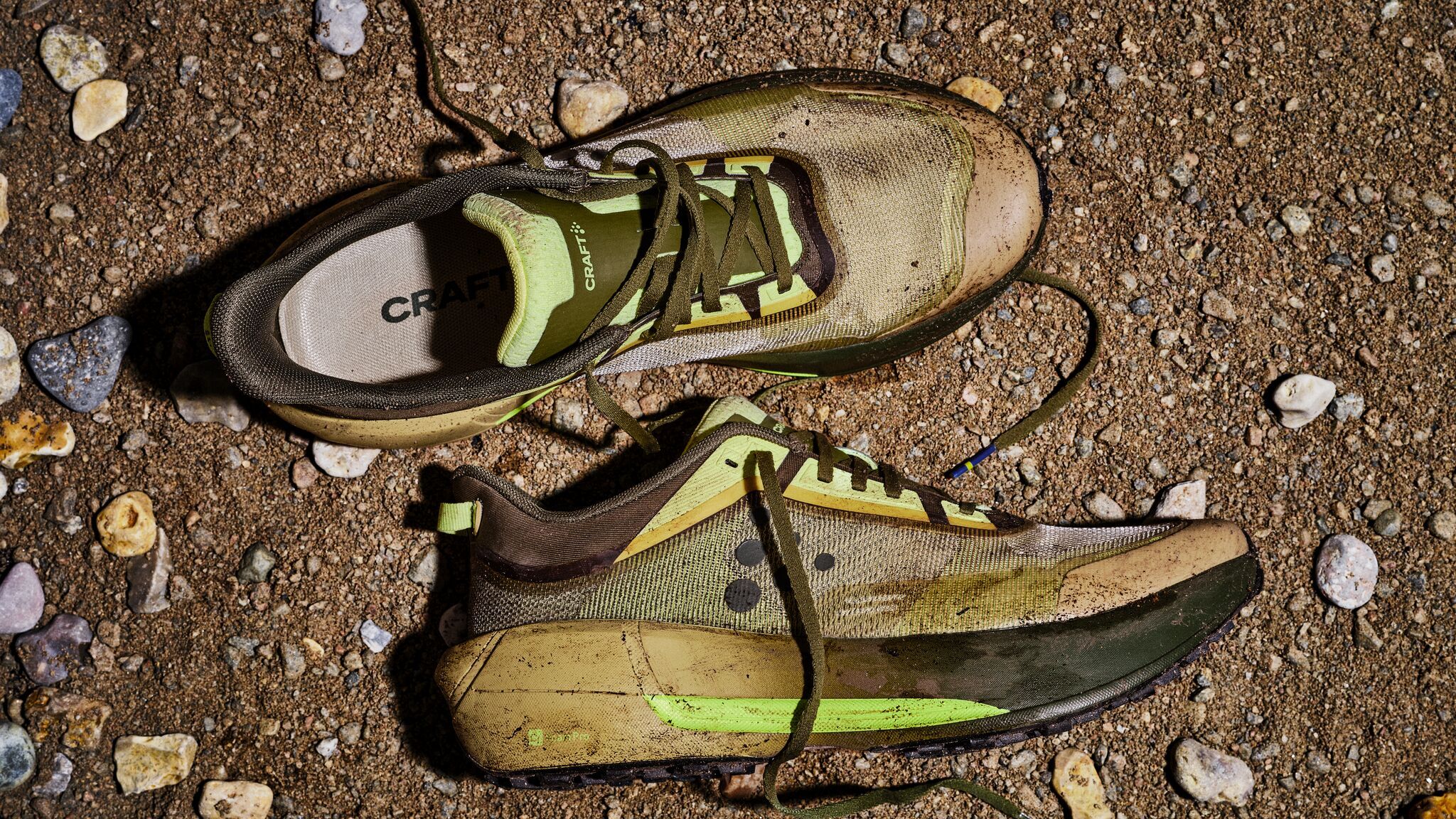 Slitte joggesko med grønne detaljer på en grusete jordoverflate, omgitt av små steiner.