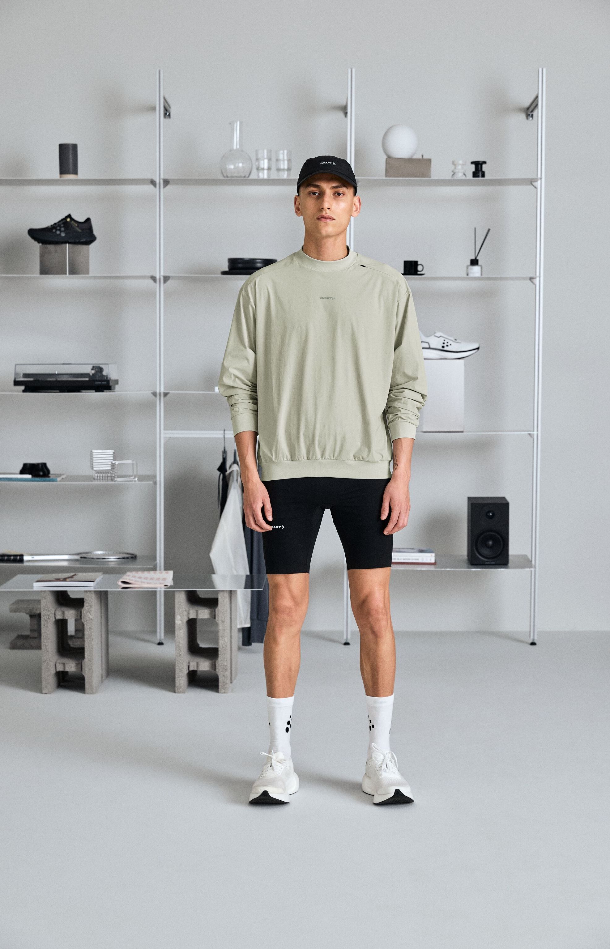 En mann i en lys genser, svarte shorts og en cap står i et minimalistisk rom med hyller fylt med pyntegjenstander og sko.