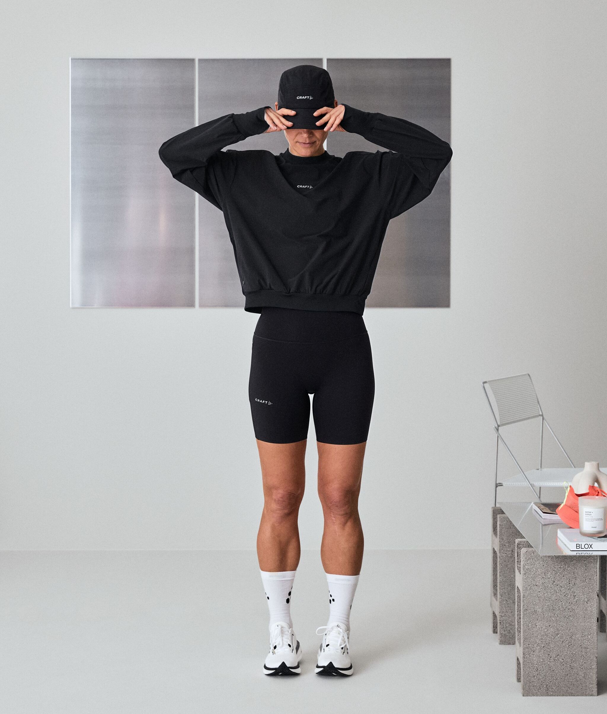 En person i svart treningsklær og cap står innendørs og retter på capen. Hvite sokker og joggesko. Minimalistisk innredning med moderne stol og bord.