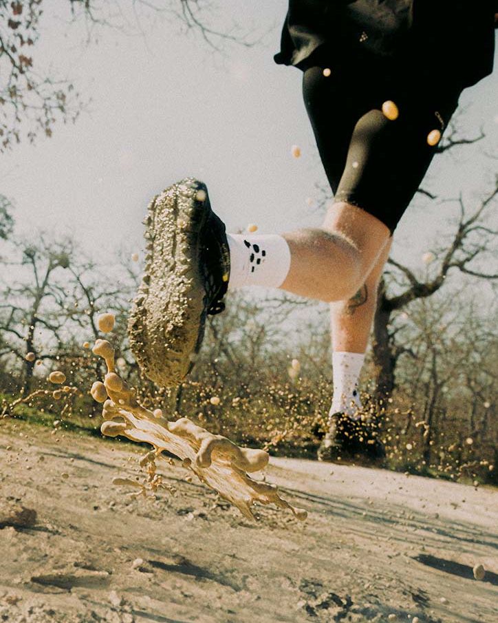 Person som løper på en gjørmete sti, spruter vann og gjørme, iført svarte sko, svarte shorts og hvite sokker. Bare trær i bakgrunnen.