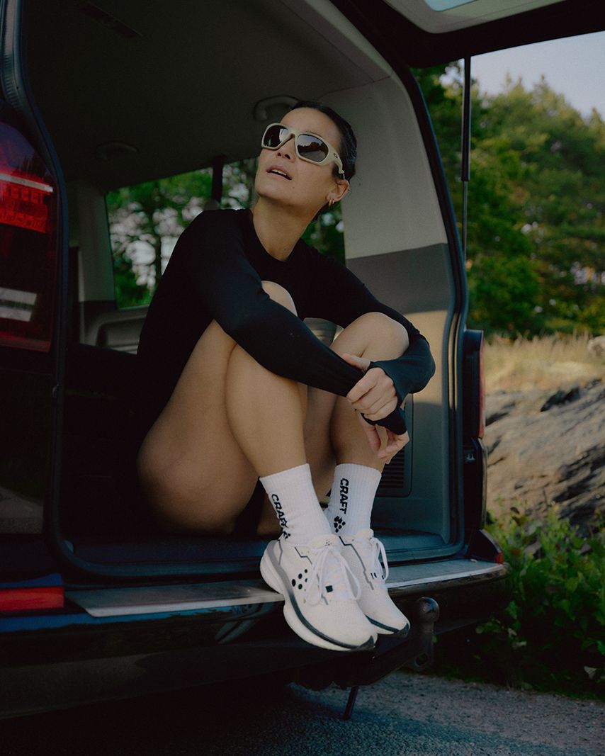 A woman in sunglasses sits in the back of a vehicle, wearing a black outfit and white sneakers, with an open landscape in the background.