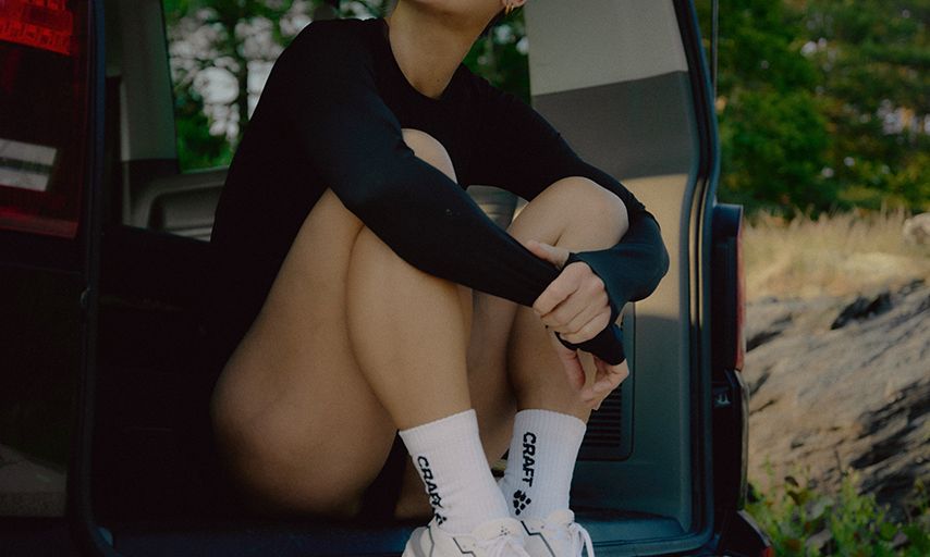 A woman in sunglasses sits in the back of a vehicle, wearing a black outfit and white sneakers, with an open landscape in the background.