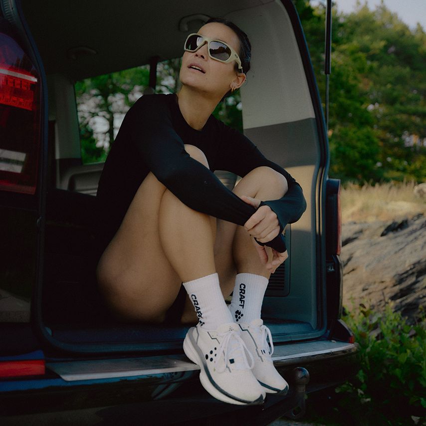A woman in sunglasses sits in the back of a vehicle, wearing a black outfit and white sneakers, with an open landscape in the background.