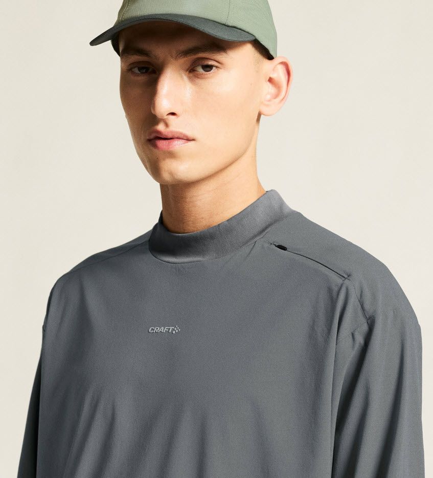 En person kledd i en grå langermet trøye med «CRAFT»-logo og en grønn baseballcap, som står foran en nøytral bakgrunn.