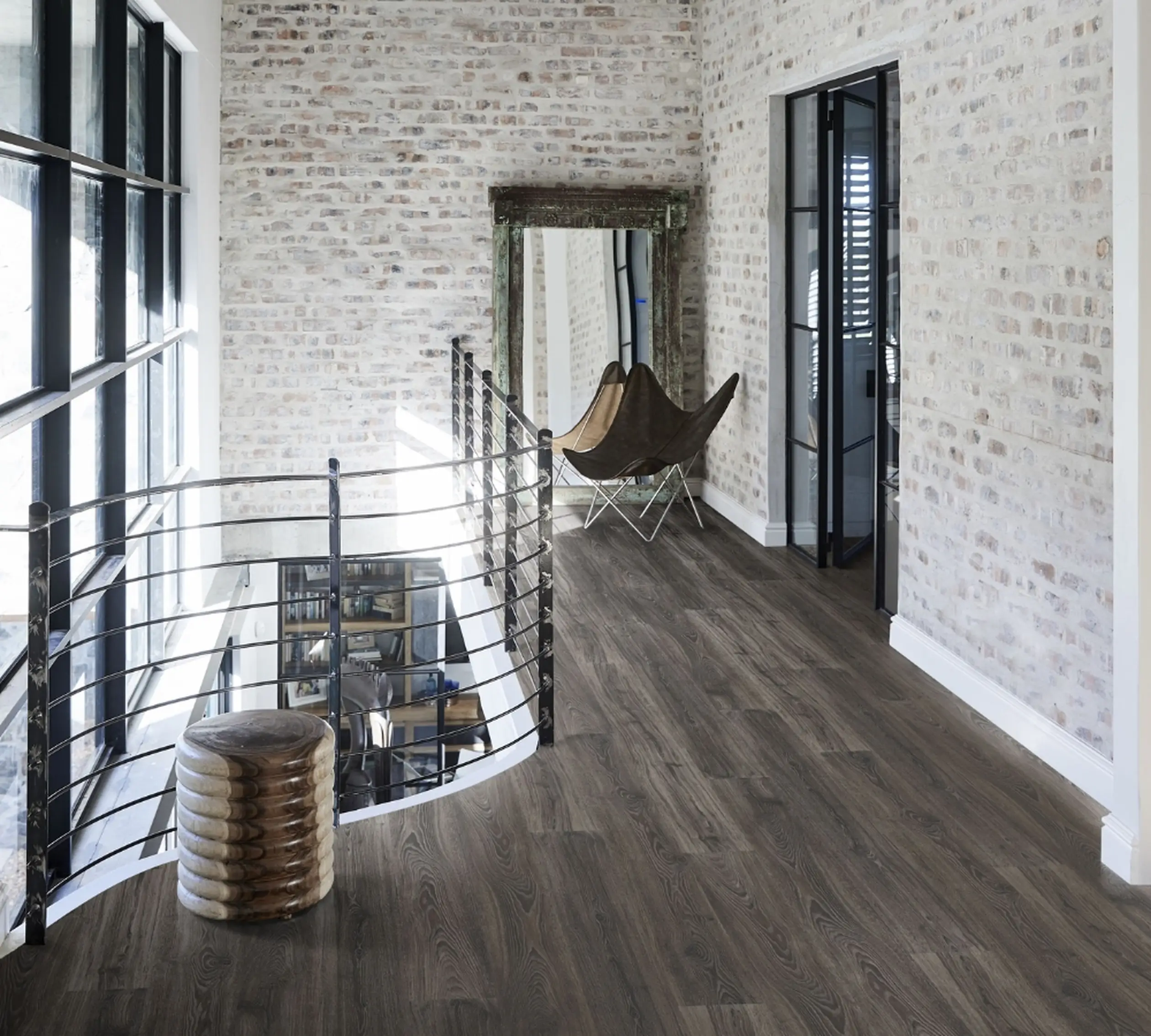 A stylish hallway with dark wood flooring, exposed brick walls, and a rustic decor piece, enhanced by natural light streaming through large windows.