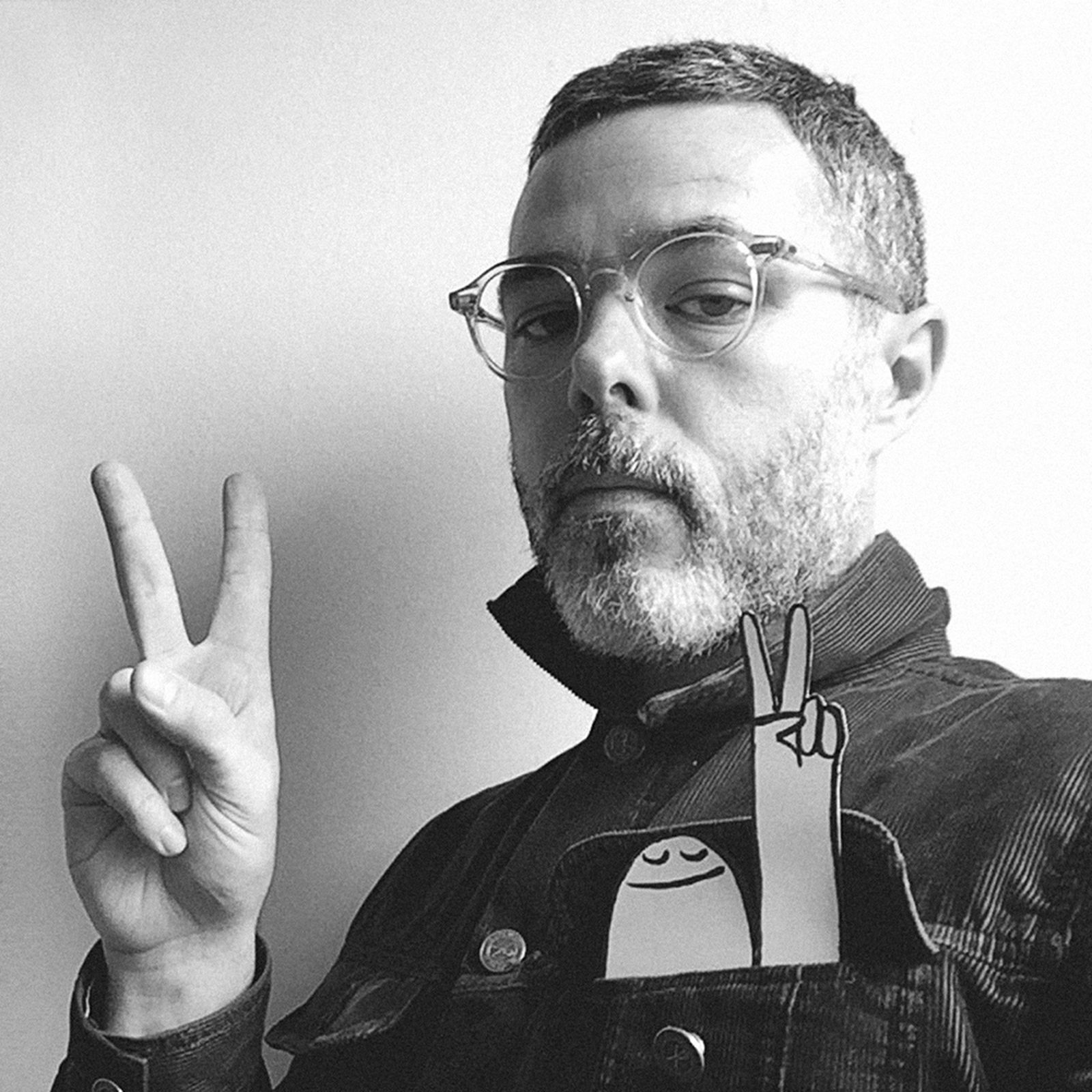Man with glasses and a beard giving a peace sign, with a cartoon character in his pocket also showing a peace sign. Black and white photo.