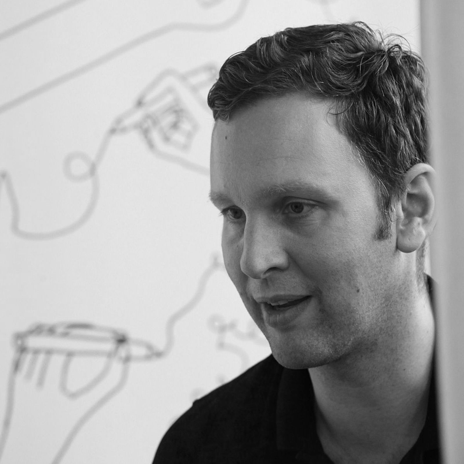 Man with short hair in a black shirt, speaking or listening, with a line drawing of hands on a white wall in the background. Black and white image.