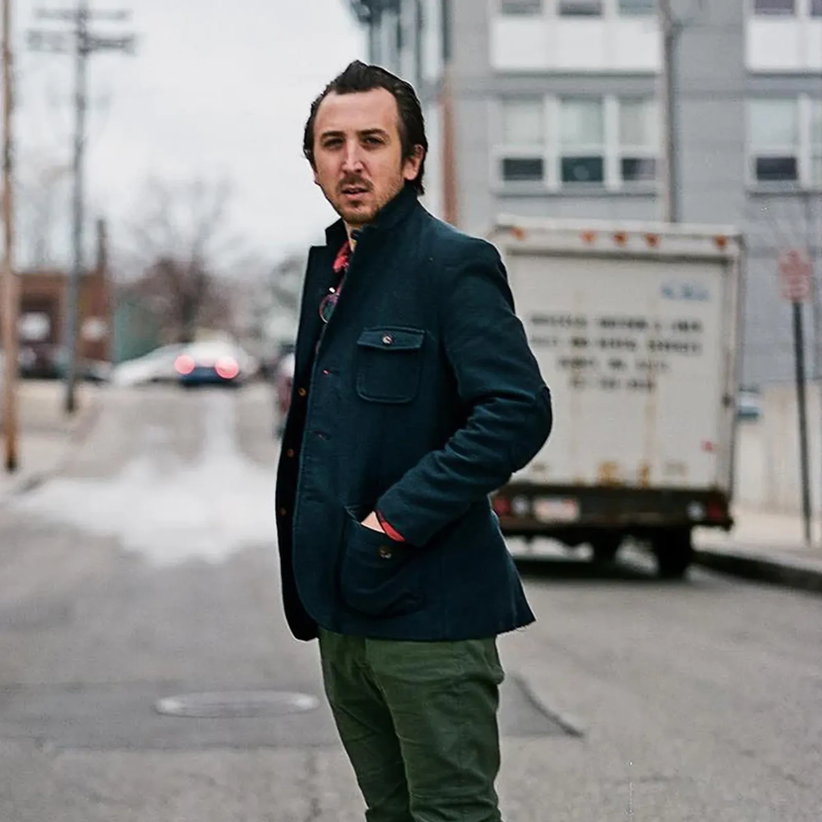 Man in a dark jacket and green pants stands on an urban street, with buildings and a truck in the background.