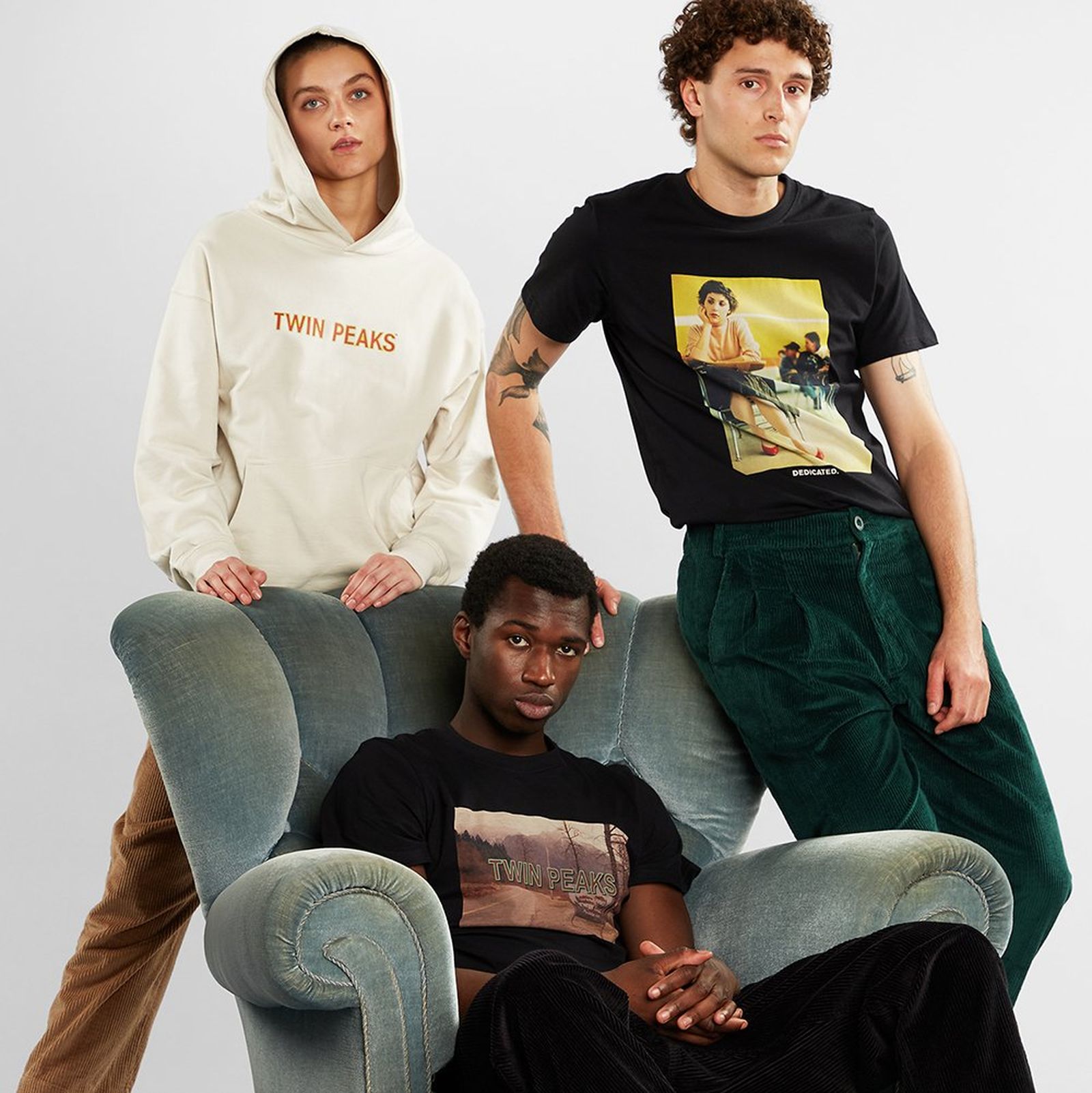 Three people in casual clothing, two standing and one sitting on a green armchair. They wear graphic shirts and a hoodie. Neutral background.