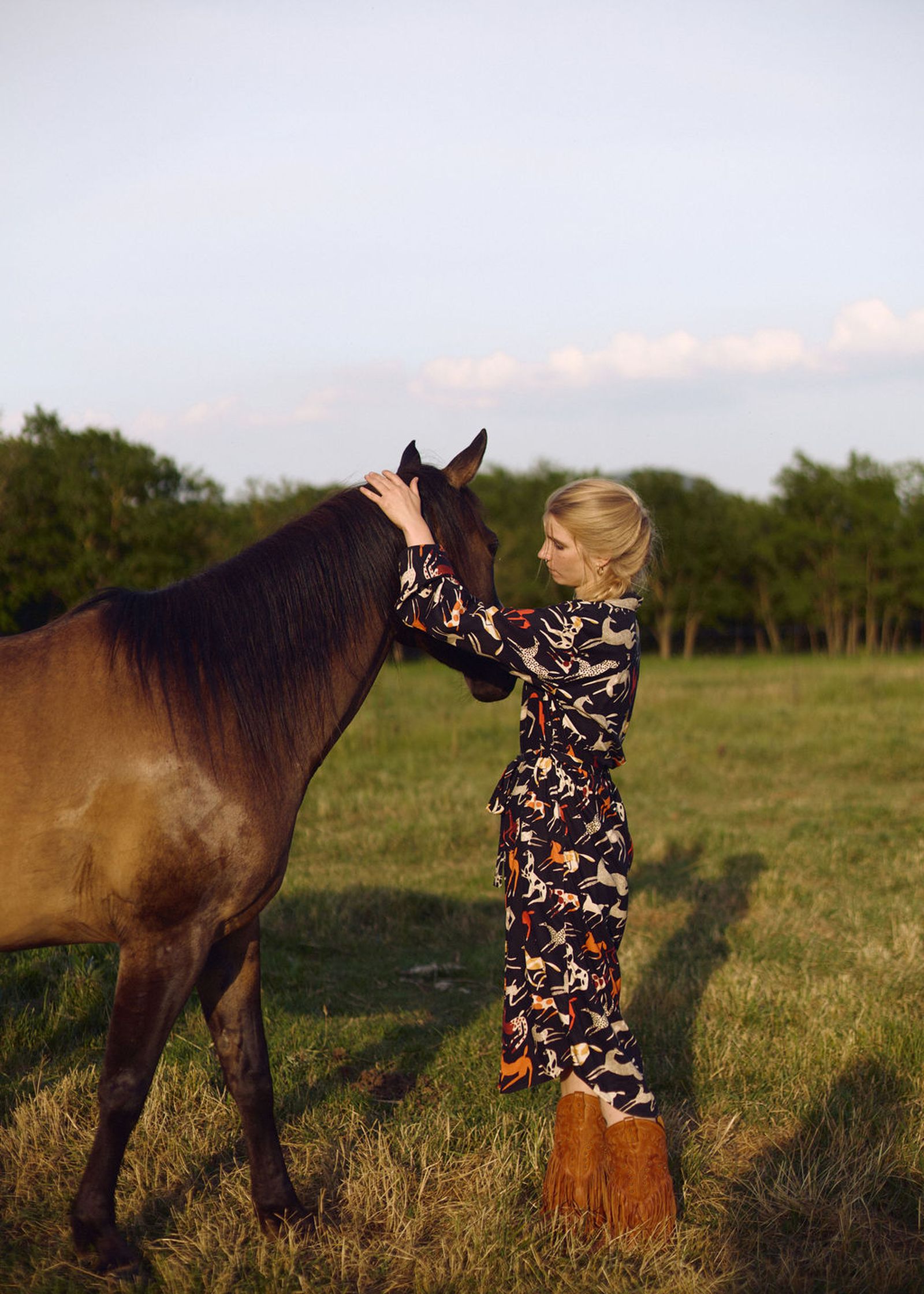 Woman in a patterned dress and brown boots gently touches a horse in a grassy field with trees in the background.