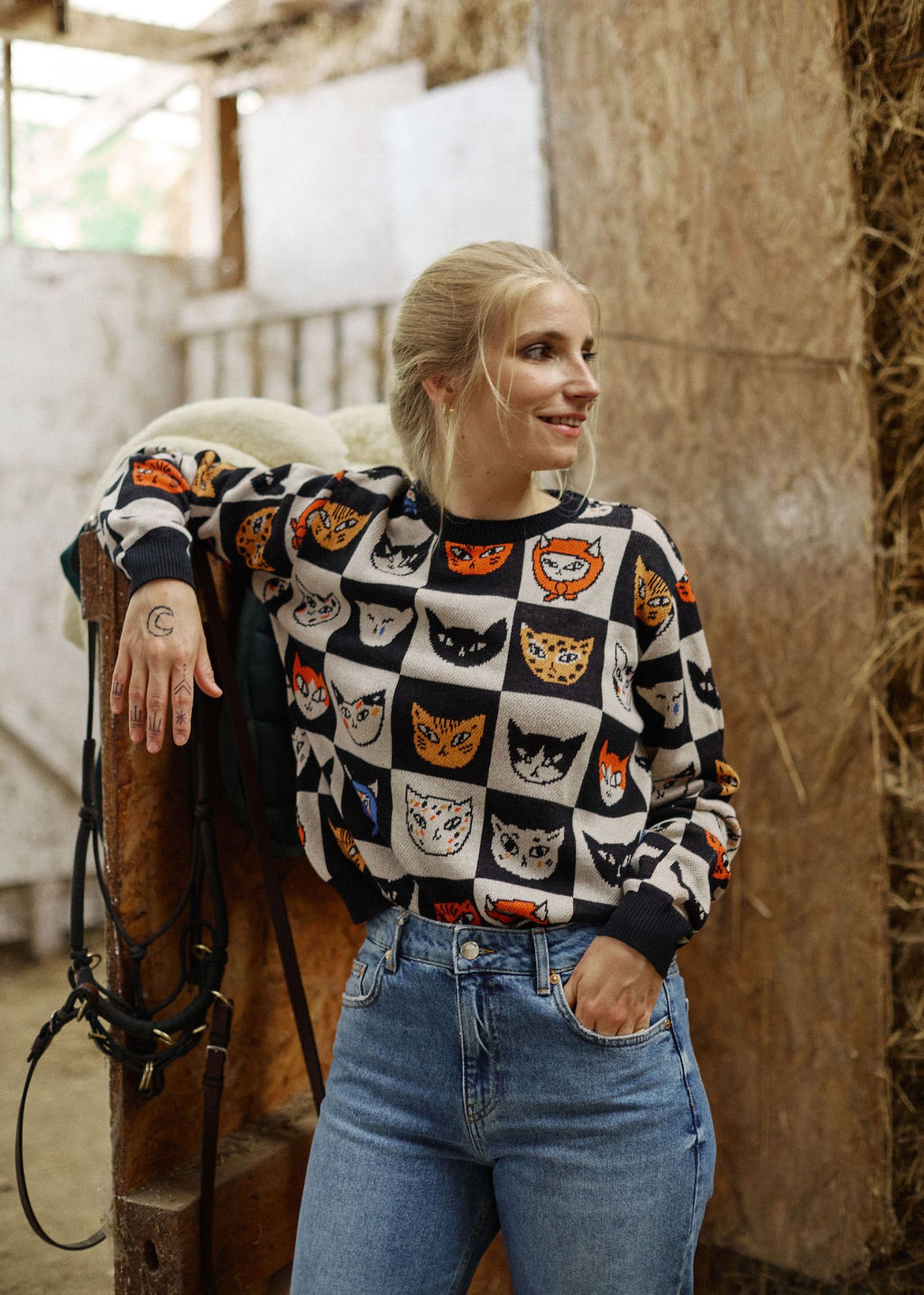 Woman in a cat-patterned sweater and jeans stands in a rustic setting, smiling with one hand in her pocket and the other resting on wooden railing.