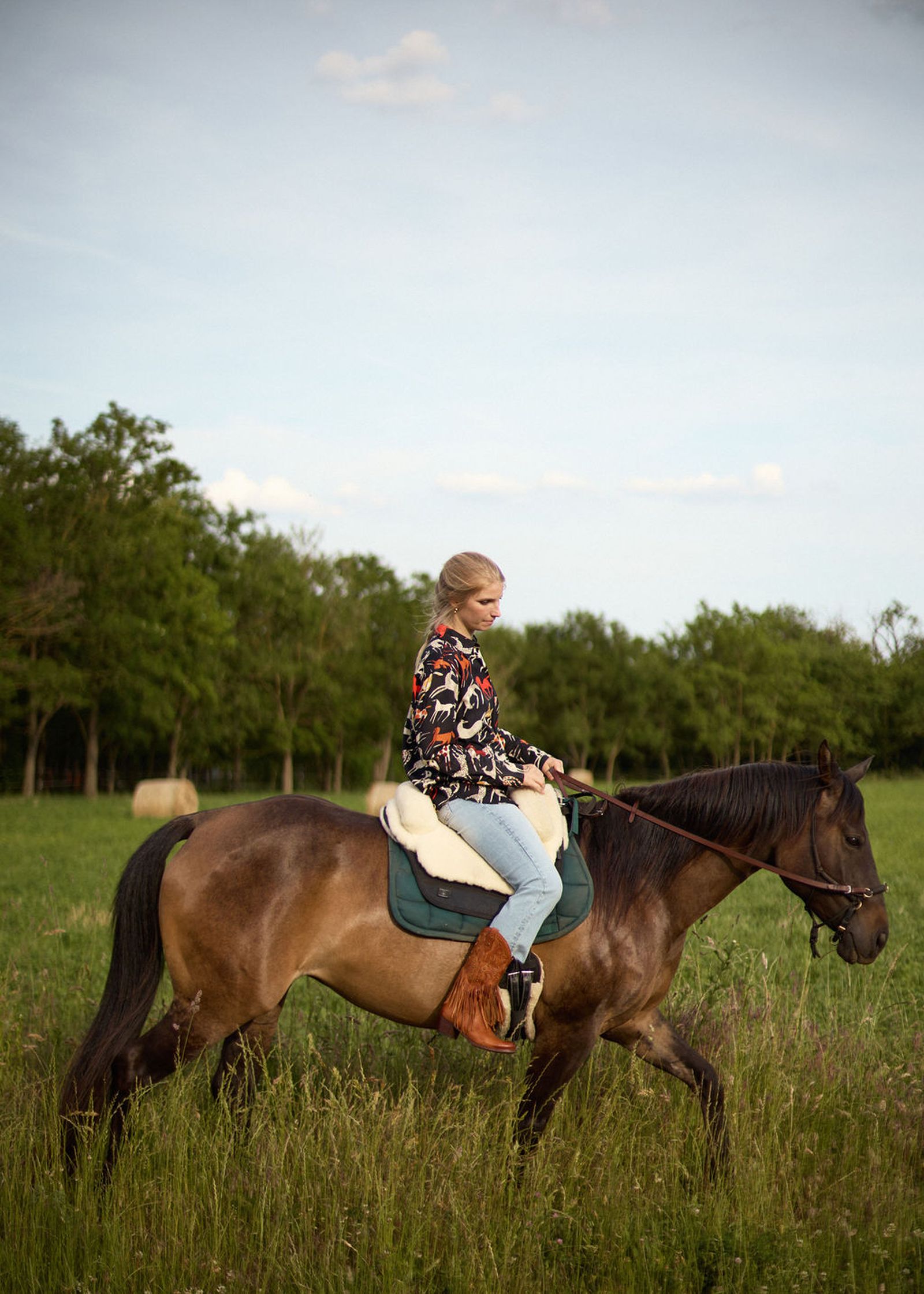 Person riding a horse in a grassy field, wearing a patterned jacket and jeans, with trees and hay bales in the background.