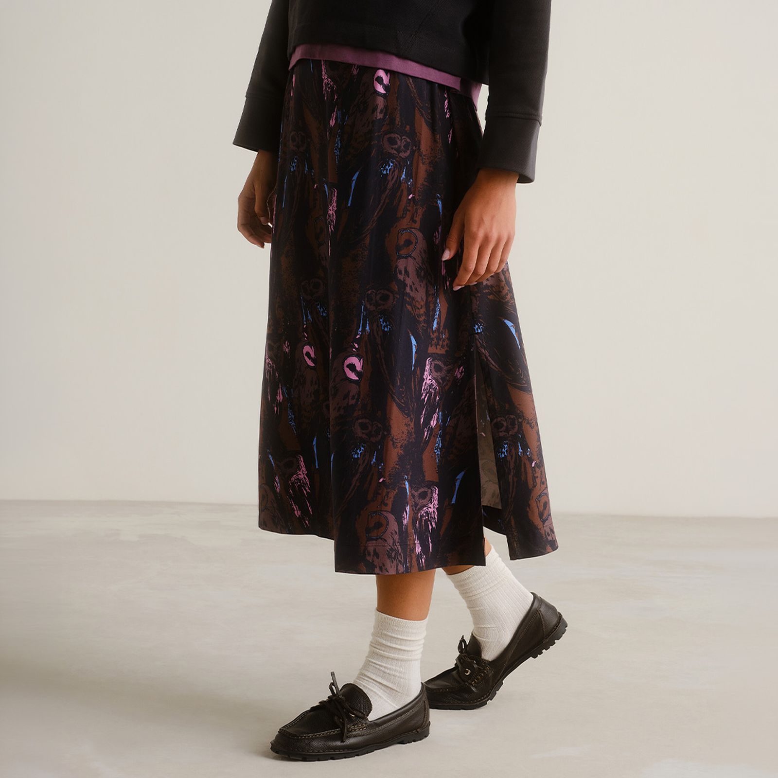 Person wearing a dark patterned skirt, white socks, and black shoes, standing on a light floor against a neutral background.