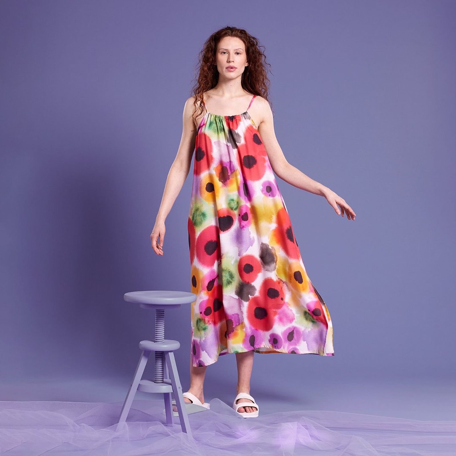 A woman in a colorful floral dress stands beside a small purple stool against a purple background.
