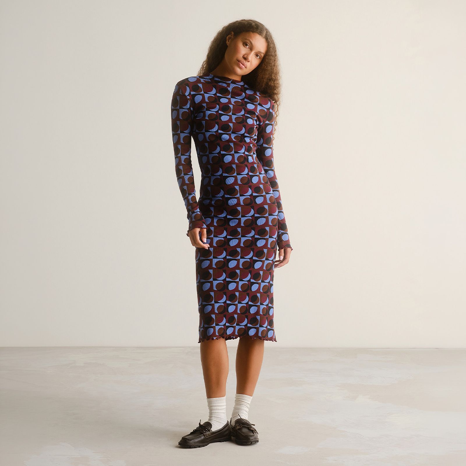 Woman in a long-sleeve, geometric patterned dress stands confidently in a minimalistic room. She's wearing white socks and black shoes.