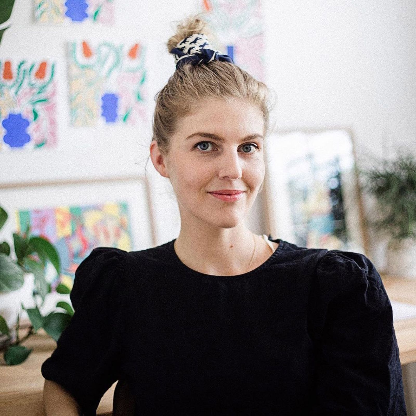 Woman with a bun and black top smiles in a room with colorful artwork and plants in the background.