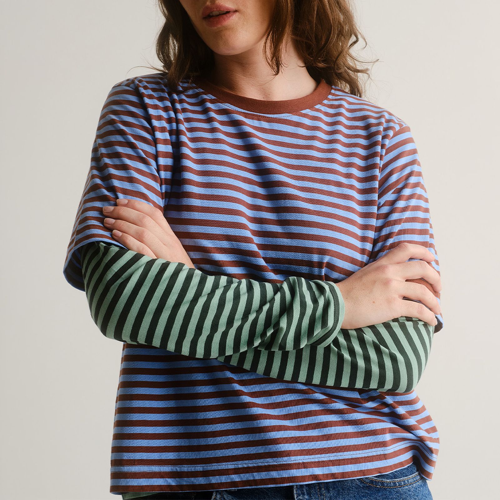 Person wearing a blue and brown striped shirt layered over a green and black striped long-sleeve shirt, arms crossed.