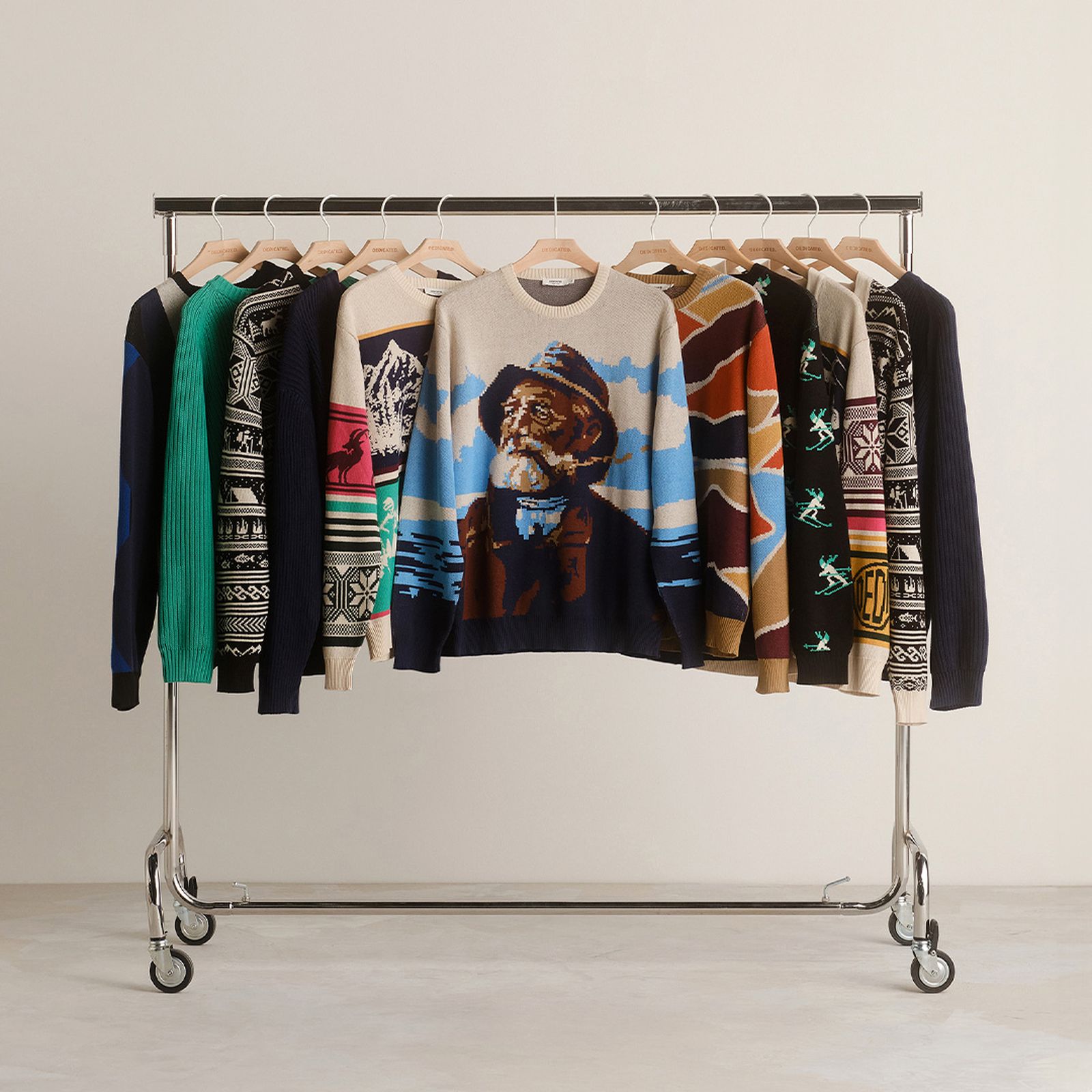 A row of colorful patterned sweaters with unique designs hangs on a metal clothing rack against a plain background.
