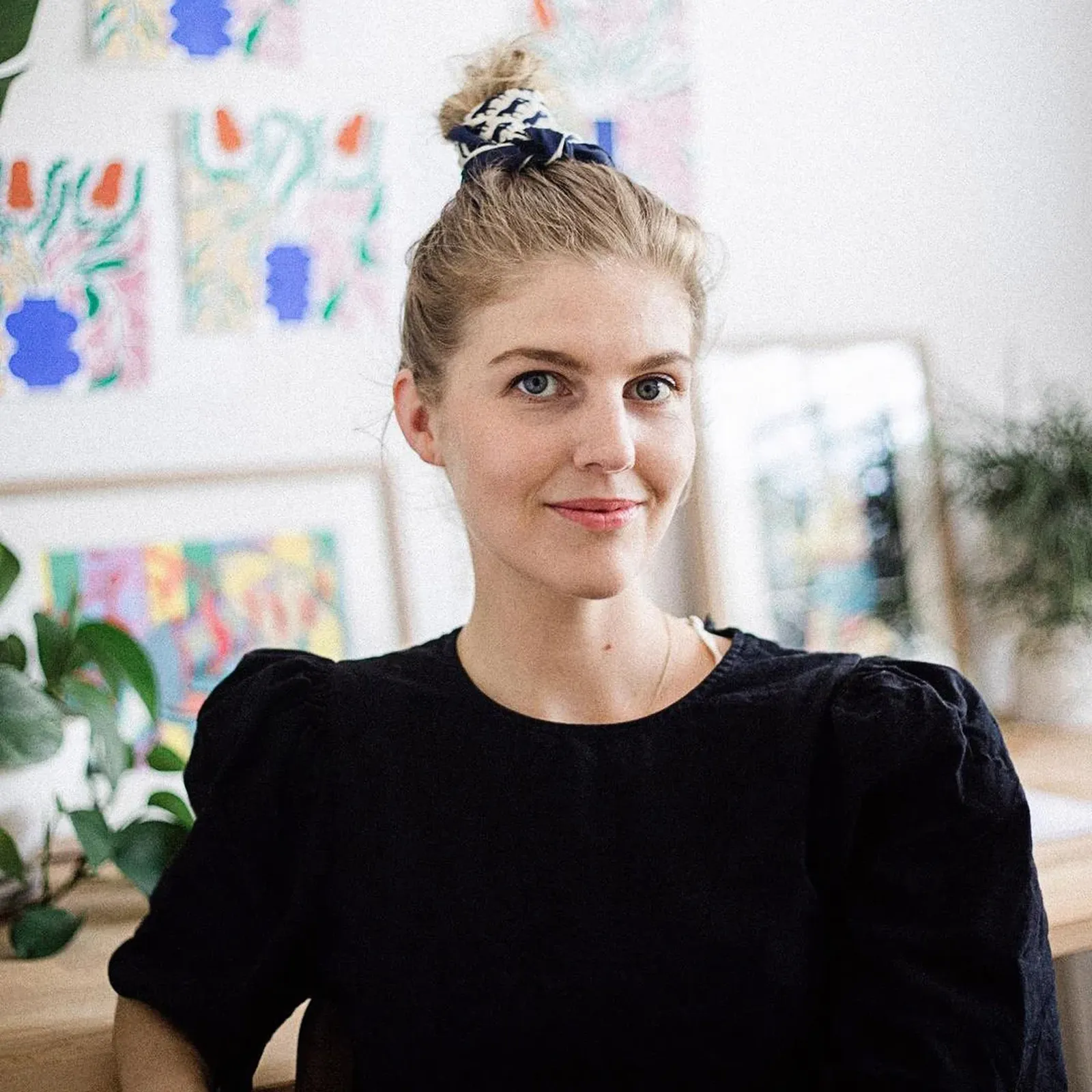 Woman with a bun, wearing a black top, sits in a bright room with colorful artwork on the wall and plants nearby.