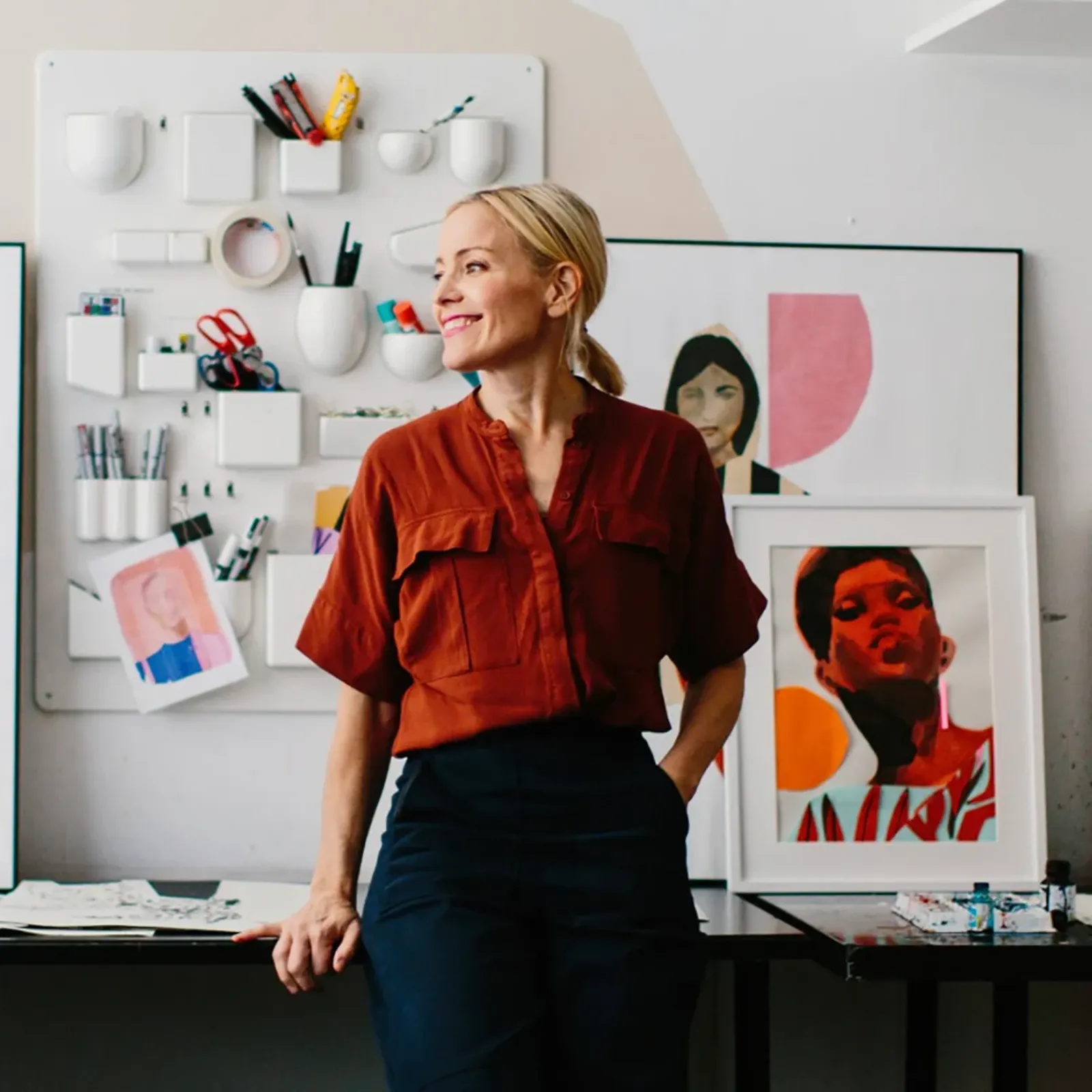 Smiling woman in a red shirt leans against a table in an art studio, surrounded by colorful portraits and art supplies on the wall.