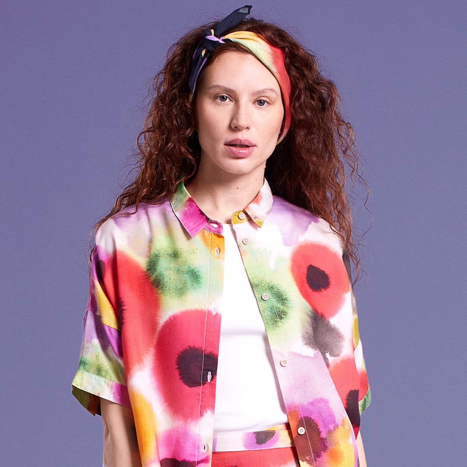 Woman with curly hair wearing a colorful, abstract-patterned outfit and headband, standing against a plain purple background.