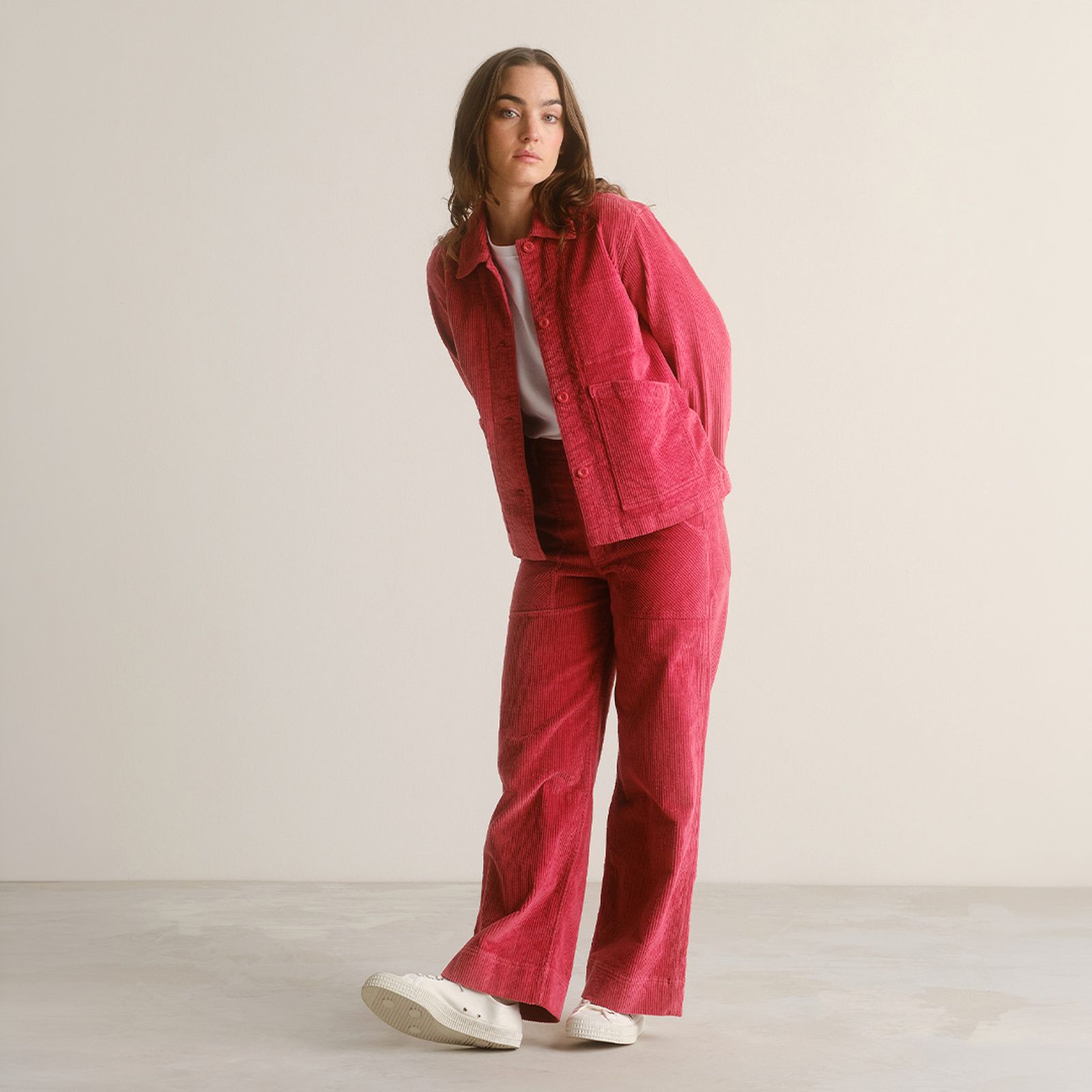A person leans slightly while wearing a bright pink corduroy suit with a white shirt and white sneakers against a plain backdrop.