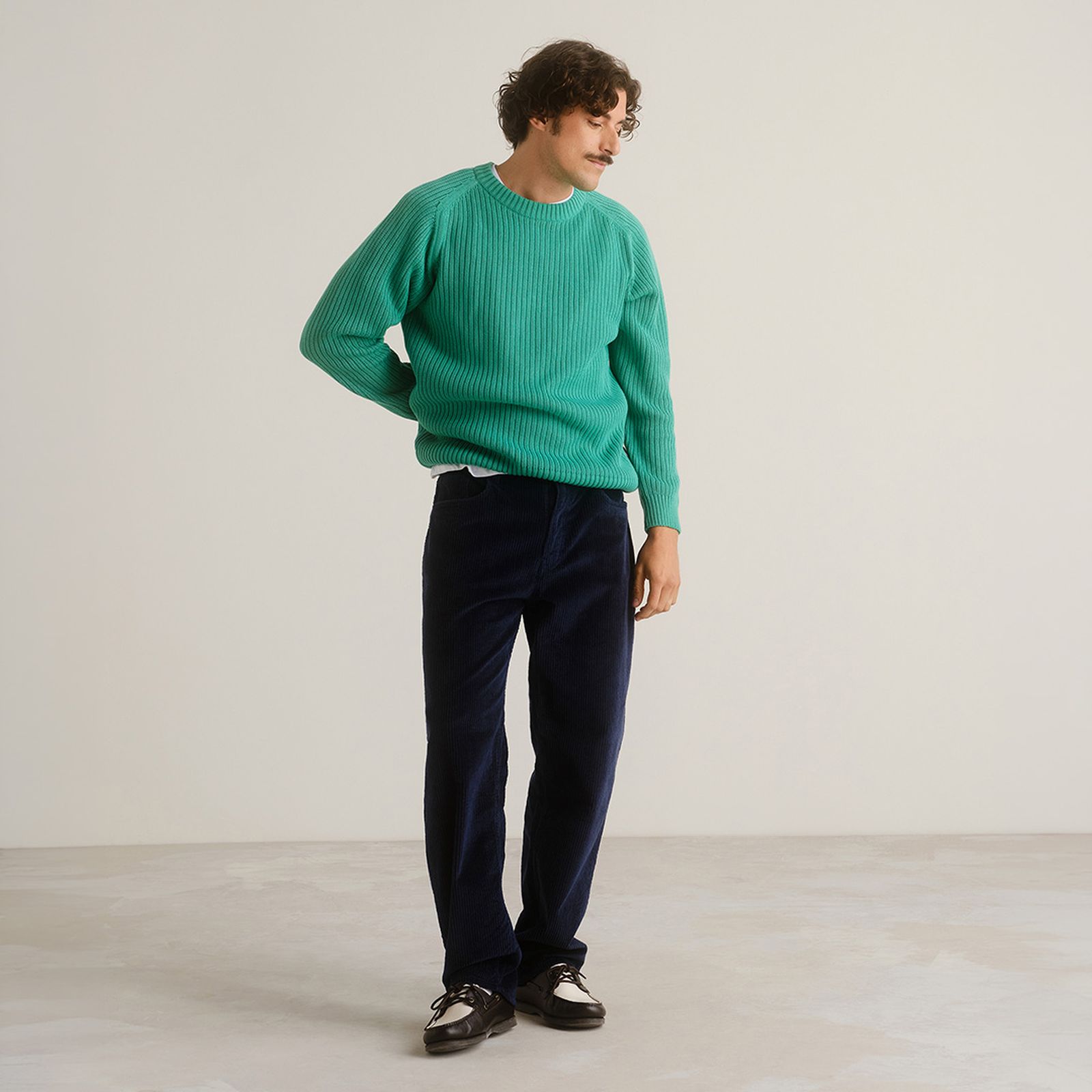 Person in a green sweater and dark pants stands on a light-colored floor against a plain background, looking to the side.