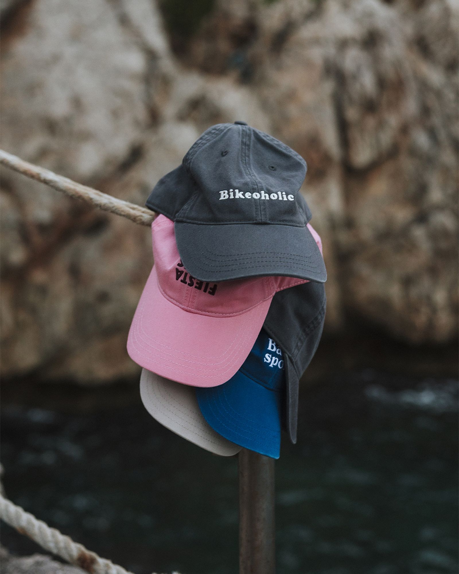 Four stacked baseball caps hanging on a rope by the sea. The top cap reads "Bikeoholic." Rocky background with water visible below.