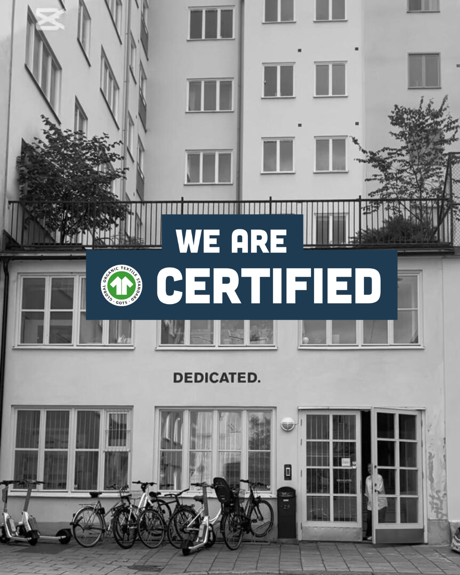 Black and white image of a building with bicycles in front. A sign reads "We are certified" with a certification logo above "DEDICATED."