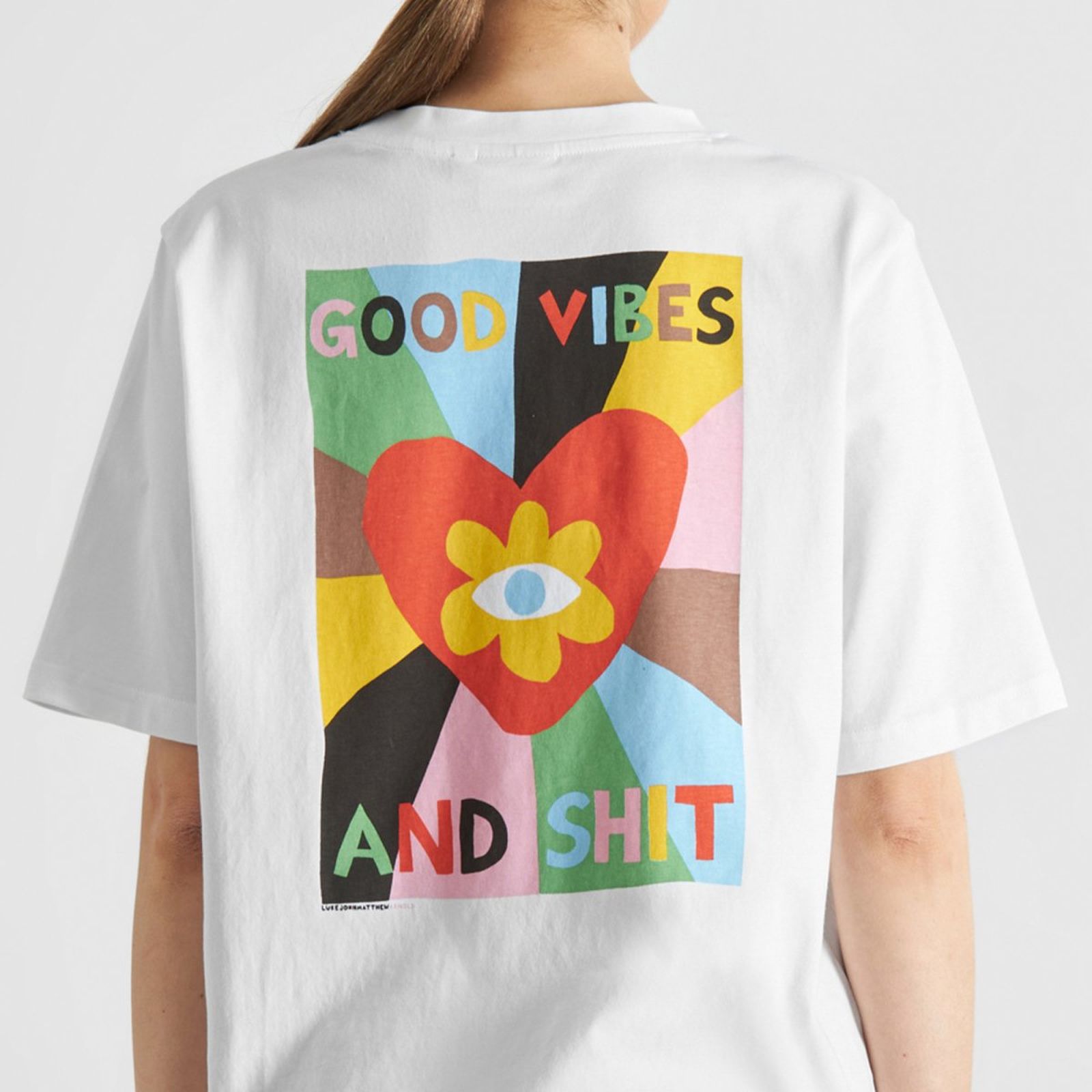 Person wearing a white T-shirt with colorful graphic of a heart, eye, and flower, featuring the text "GOOD VIBES AND SHIT."