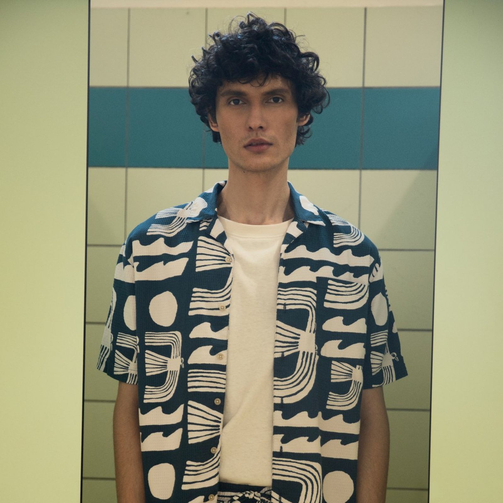 Person with curly hair wearing a patterned shirt stands in front of a tiled wall with green and blue accents.