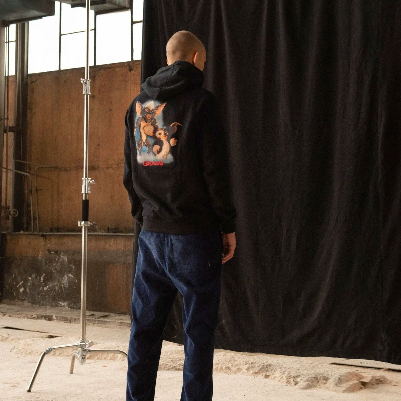 Person in a black hoodie with a cartoon graphic on the back, standing in a studio with industrial decor and a black curtain backdrop.