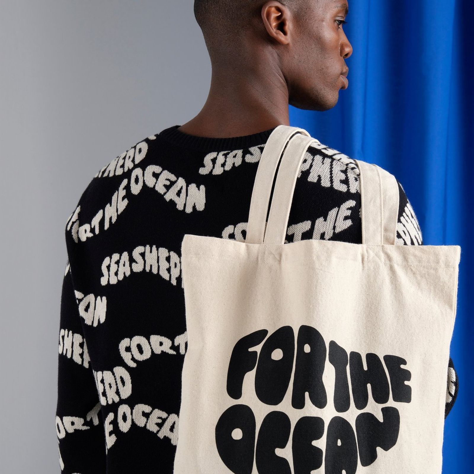Man wearing a sweater with "Sea Shepherd" text, holding a tote bag with "For the Ocean" text, standing against a blue and gray background.