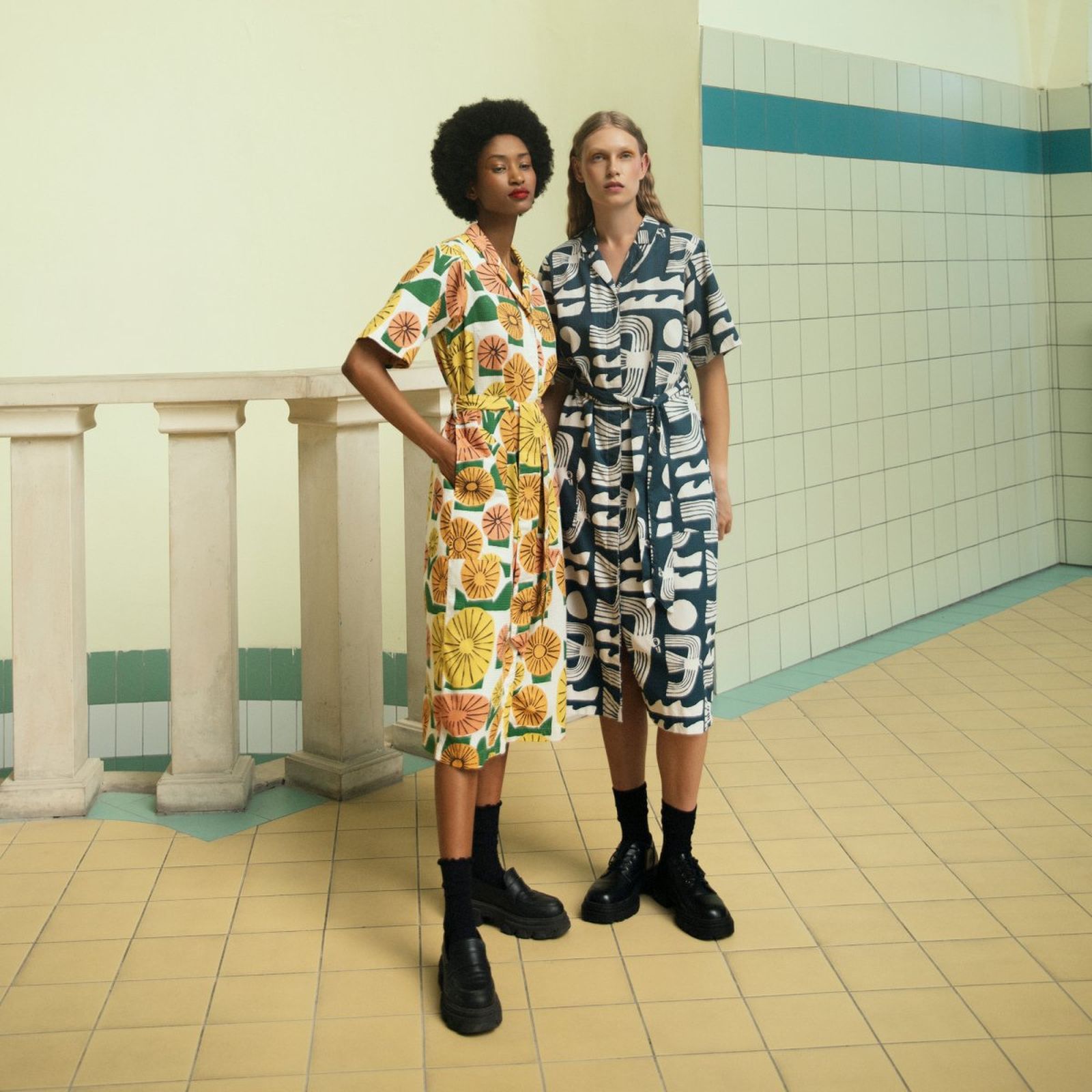 Two models in patterned dresses and black boots stand in a tiled room with pale yellow floors and light green walls.