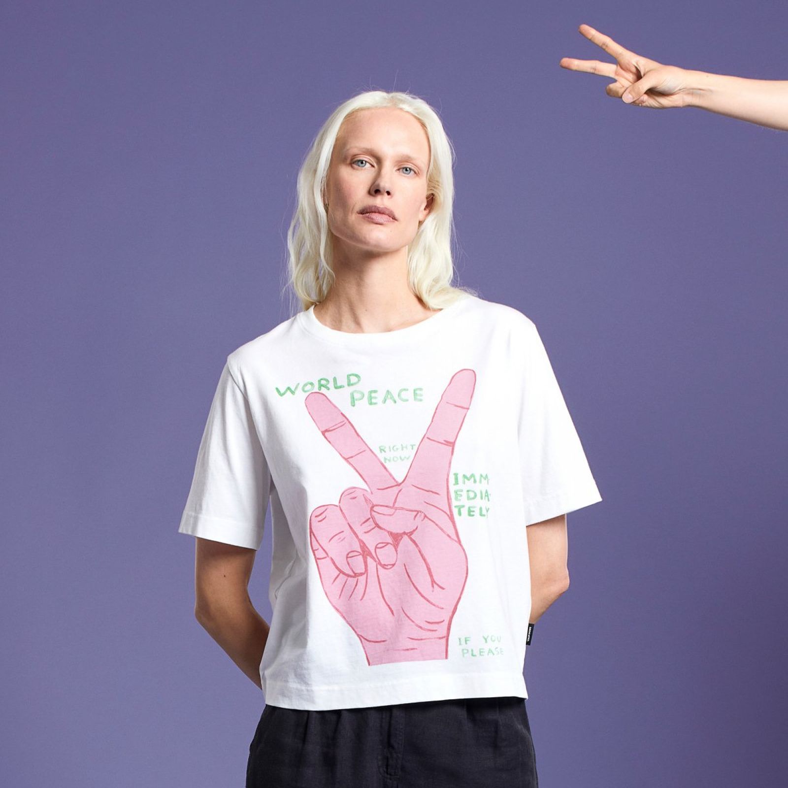 Person with long blonde hair in a "World Peace" T-shirt stands against a purple background. A hand making a peace sign is visible on the right.